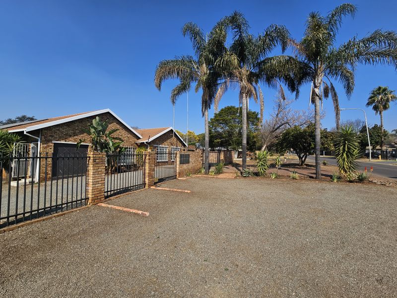 Front exterior view of the office property featuring a gravel parking area, brick facade, tiled roof, and secure perimeter fencing with mature palm trees.