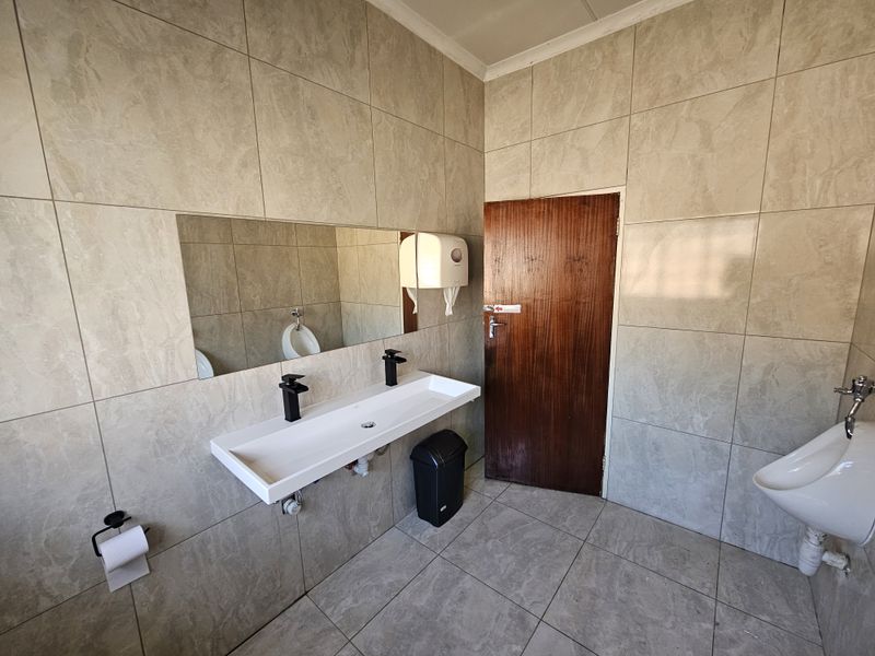 A clean, modern office bathroom featuring full-height wall tiling, a wall-mounted basin with black fixtures, and a wall-mounted urinal.