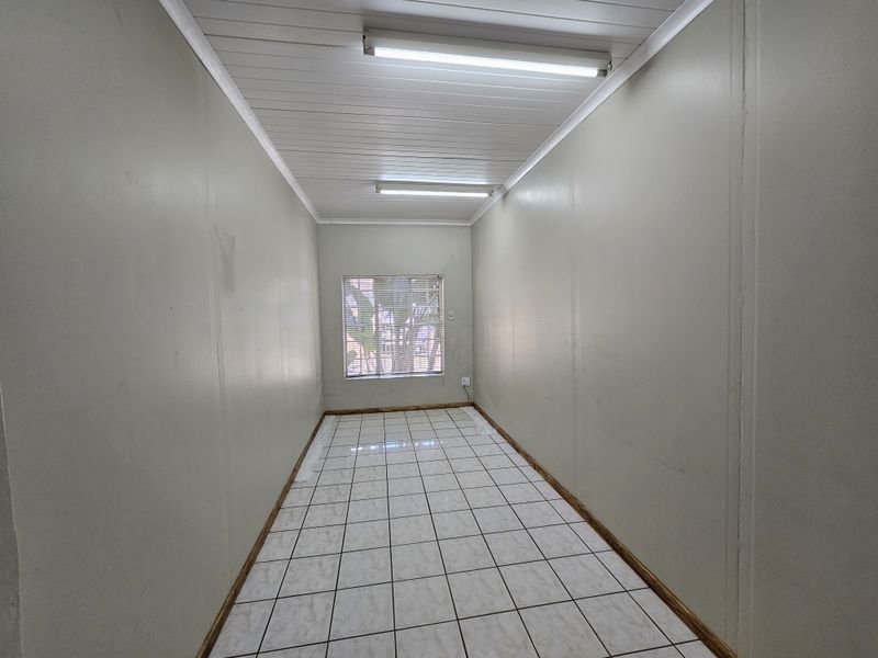 A vacant, rectangular office space featuring tiled flooring, fluorescent lighting, and a window with blinds.