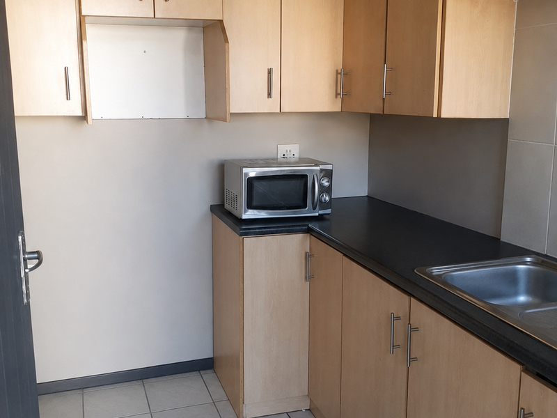 A compact kitchen area featuring light wood-tone cabinetry, dark laminate countertops, a stainless steel sink, and neutral tiled flooring.