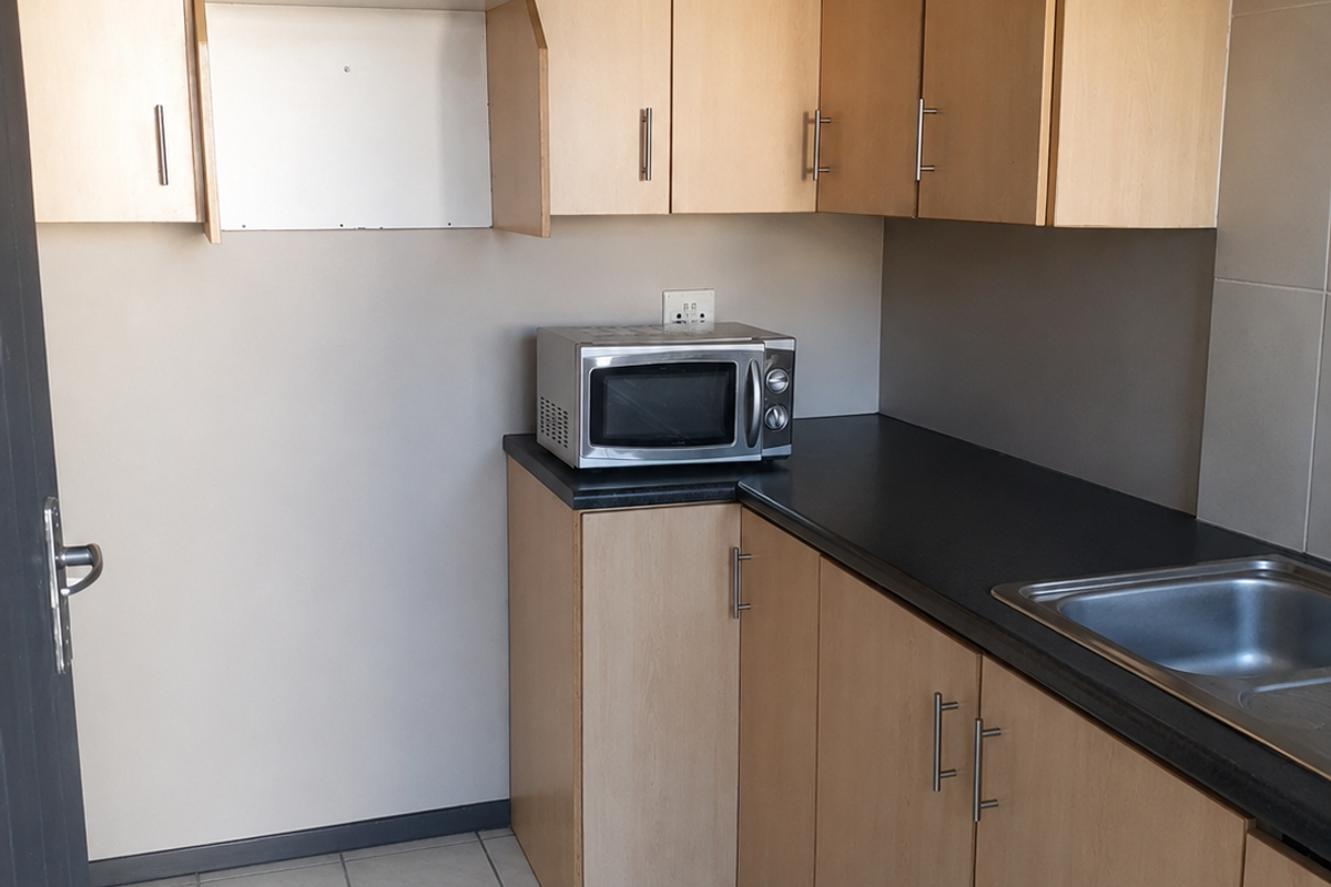 A compact kitchen area featuring light wood-tone cabinetry, dark laminate countertops, a stainless steel sink, and neutral tiled flooring.