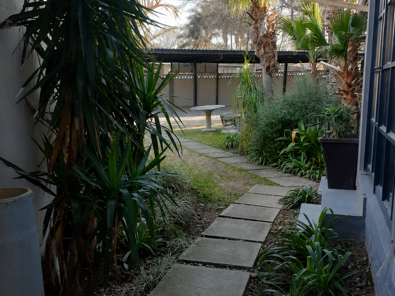 A rear garden pathway featuring concrete pavers leading towards a covered parking area, bordered by established landscaping and building exterior.