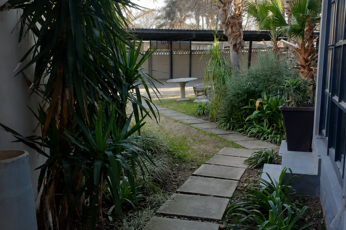 A rear garden pathway featuring concrete pavers leading towards a covered parking area, bordered by established landscaping and building exterior.