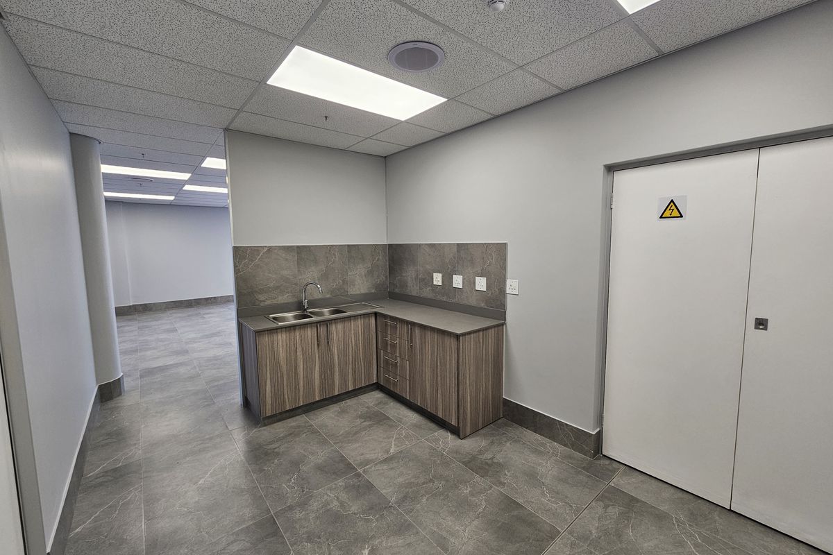 A clean, modern kitchenette area featuring wood-look cabinetry, a stainless steel sink, stone-effect floor tiles, and a dedicated electrical cupboard in an industrial space.
