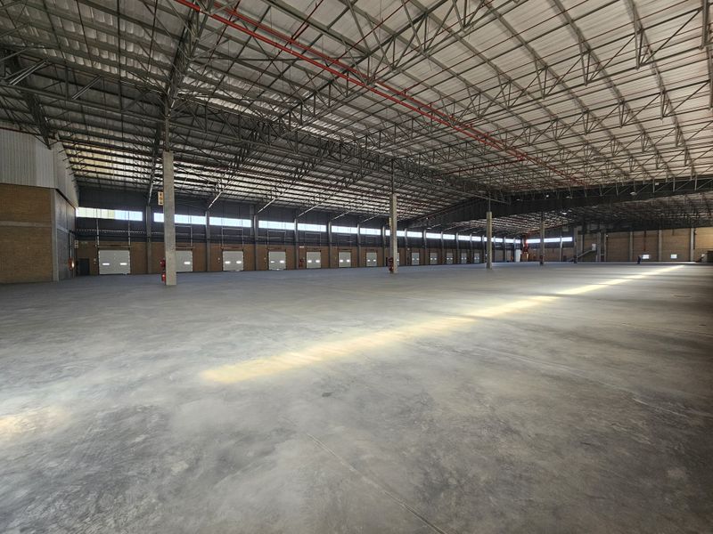 Expansive industrial warehouse space featuring high-volume exposed steel roof trusses, polished concrete flooring, and multiple roller shutter doors for efficient logistics access.