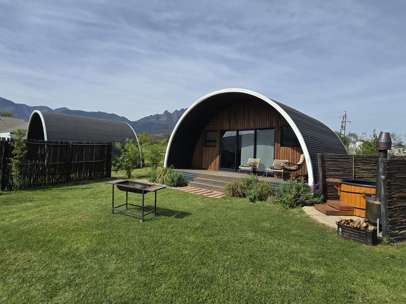 Modern curved-roof cabin featuring timber cladding, a private wooden deck, and an outdoor wood-fired hot tub set within a landscaped farm garden.