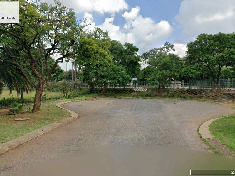 Street view of the vacant commercial land showing asphalt road access, perimeter security fencing, and established trees.