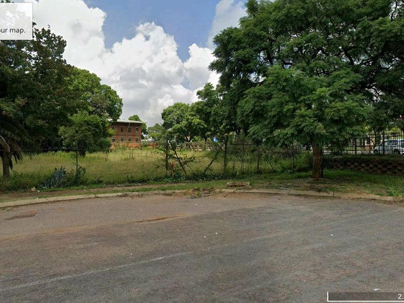 A view of a vacant commercial land plot featuring mature trees, perimeter fencing, and direct access from the paved street.