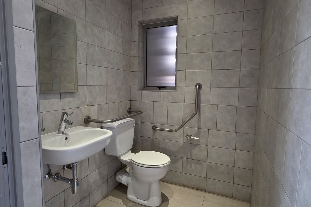 A functional, tiled bathroom featuring a wall-mounted basin, toilet, and safety grab rails, suitable for accessibility requirements.