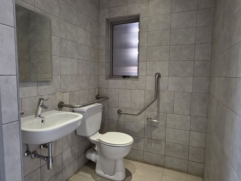 A functional, tiled bathroom featuring a wall-mounted basin, toilet, and safety grab rails, suitable for accessibility requirements.