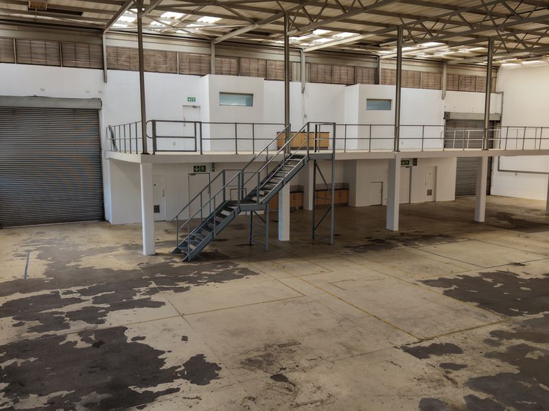 Spacious industrial warehouse featuring a mezzanine level, metal staircase, high ceilings with skylights, and large roller shutter doors for easy access.