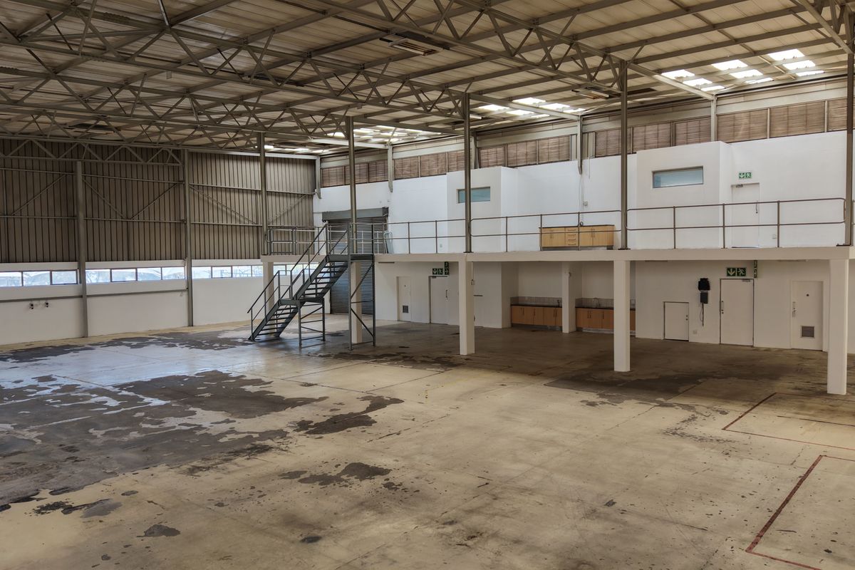 Spacious industrial warehouse featuring a high-clearance steel truss roof, mezzanine level with staircase, roller shutter access, and integrated storage cabinetry.