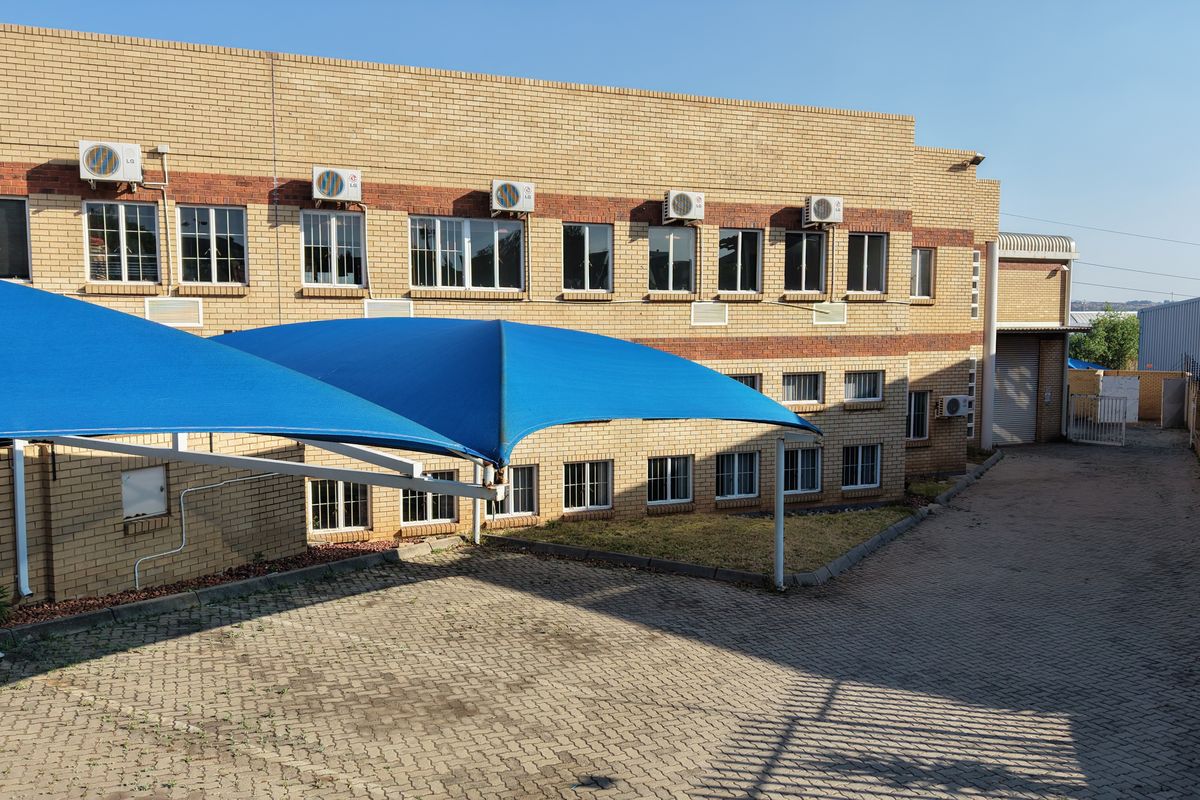 Rear exterior view of an industrial building featuring a brick facade, paved driveway, and blue shade sail parking structures.