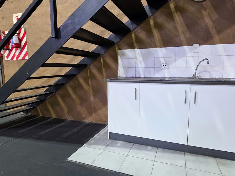 A functional utility area featuring a compact kitchenette with sink and cabinetry, situated beneath a metal staircase in an industrial space.