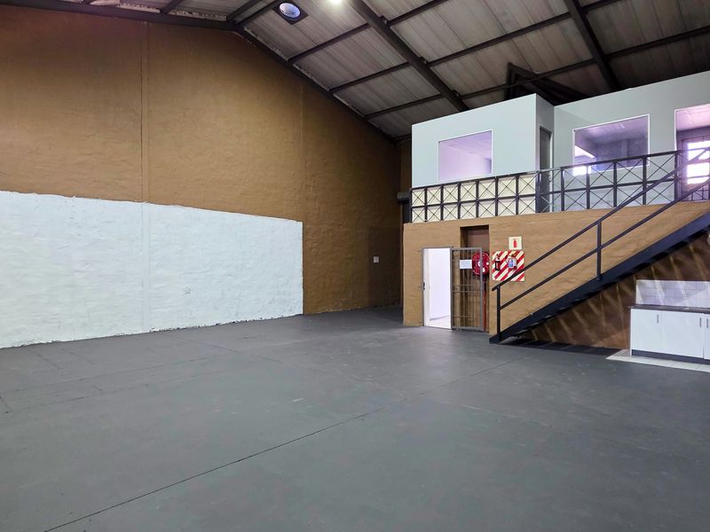 Spacious industrial warehouse unit featuring a mezzanine office level, concrete flooring, exposed steel roof trusses, and a small built-in kitchenette area.