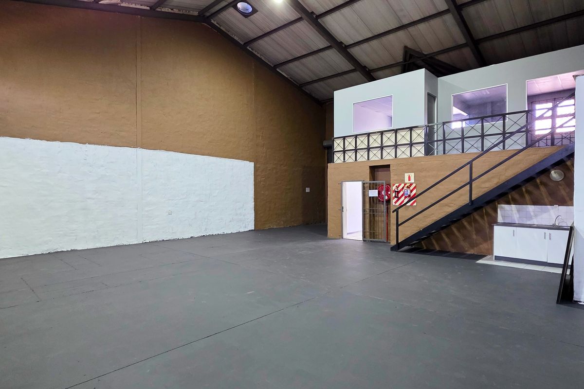 Spacious industrial warehouse unit featuring a mezzanine office level, concrete flooring, exposed steel roof trusses, and a small built-in kitchenette area.