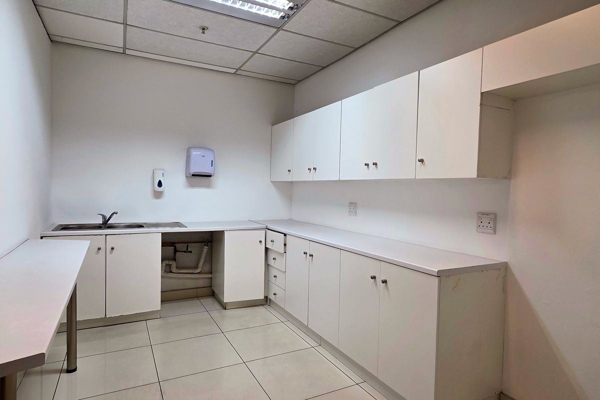 A functional industrial kitchenette featuring white built-in cabinetry, laminate countertops, a stainless steel sink, and tiled flooring with a drop ceiling.