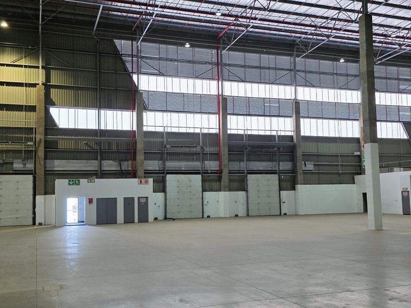 Spacious industrial warehouse featuring high ceilings, concrete flooring, multiple roller shutter doors, and exposed steel structural elements, suitable for logistics or manufacturing operations.