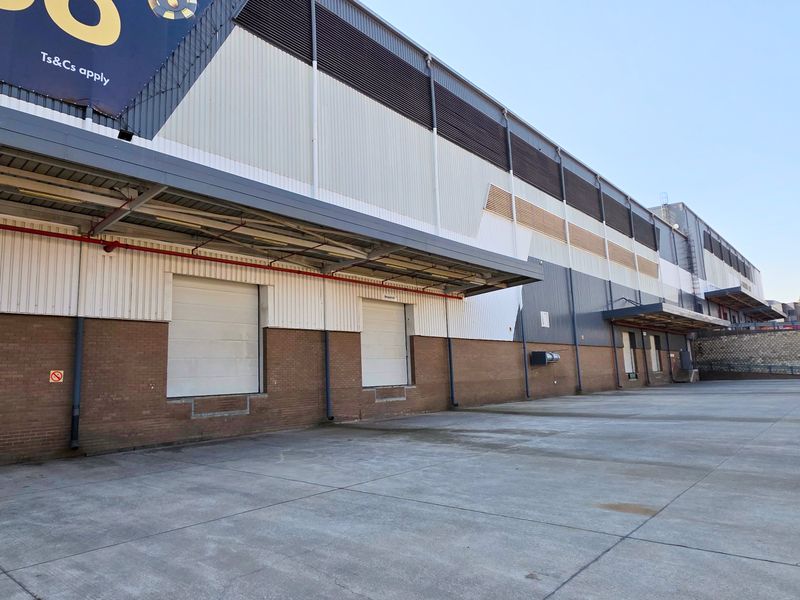 Industrial warehouse exterior featuring multiple loading bays with roller shutter doors, concrete yard space, and metal cladding facade.