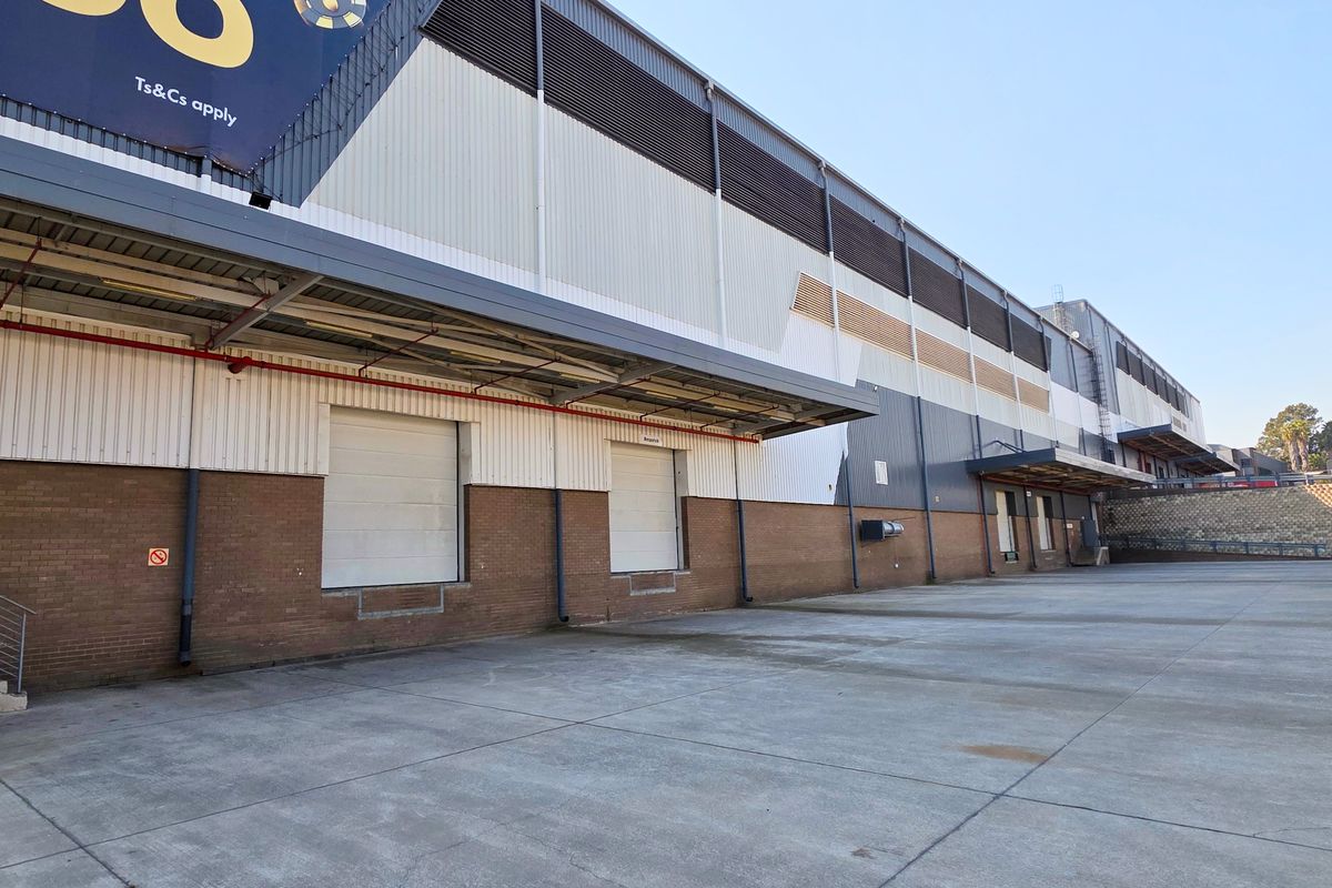Industrial warehouse exterior featuring multiple loading bays with roller shutter doors, concrete yard space, and metal cladding facade.
