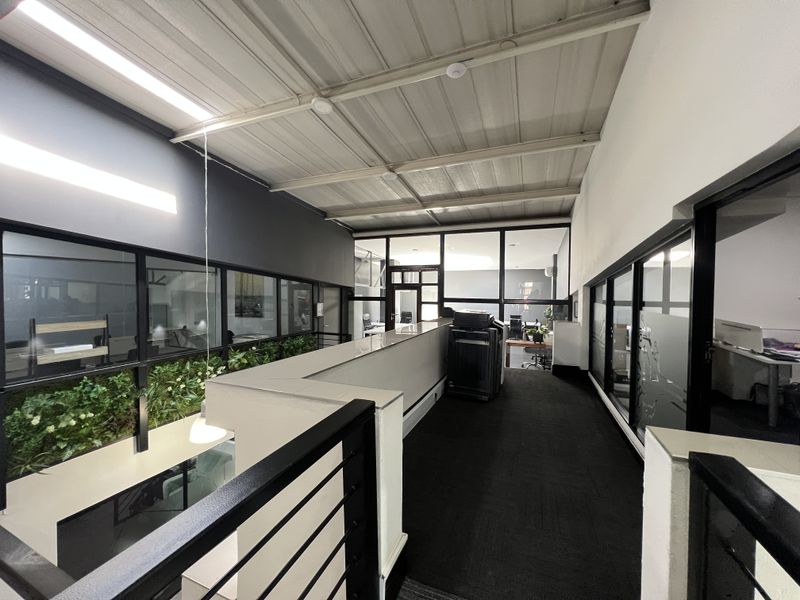 A modern office hallway featuring exposed metal ceilings, glass partition walls, and a mezzanine-style layout overlooking the lower office level.
