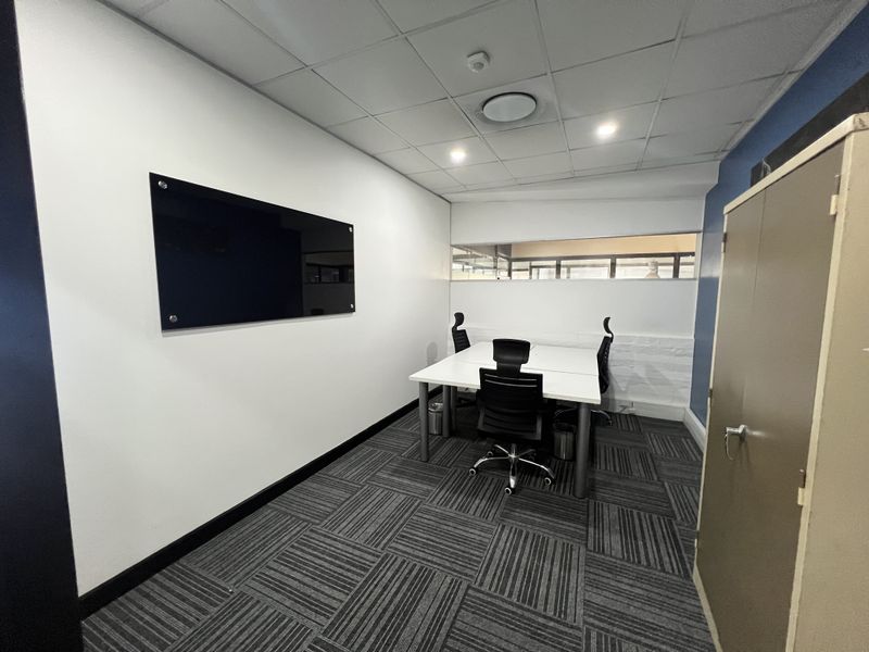 A functional office space featuring carpet tiles, a suspended ceiling with recessed lighting, a wall-mounted glass whiteboard, and built-in storage.