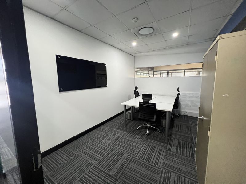 A functional office space featuring carpet tiles, a suspended ceiling with recessed lighting, and a wall-mounted glass whiteboard.