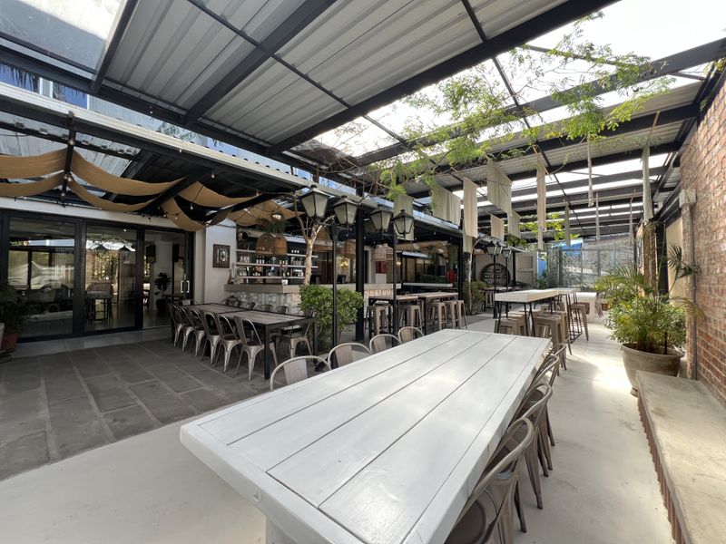 A spacious covered outdoor patio area featuring a metal roof structure, exposed brick wall, and a built-in bar counter, ideal for office social gatherings.