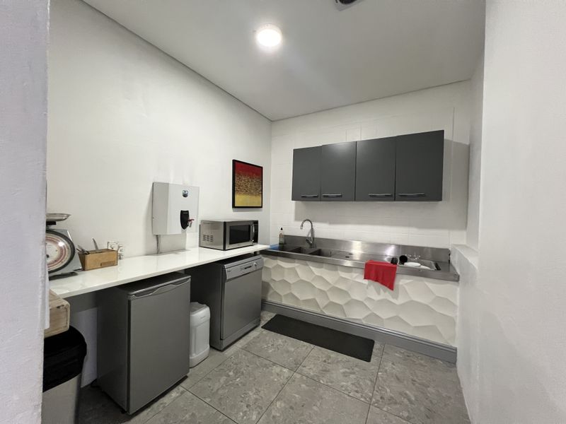 A functional office kitchenette featuring stainless steel sink, wall-mounted cabinetry, textured wall detailing, and tiled flooring.
