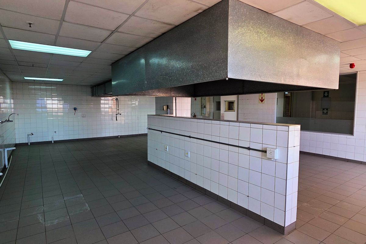 An industrial kitchen space featuring tiled surfaces, a large central extractor hood, suspended ceiling panels, and multiple plumbing and electrical connection points.