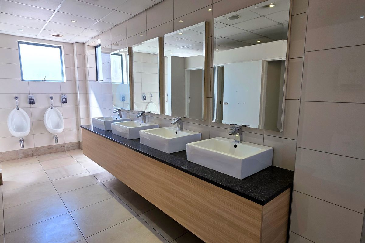A clean, modern industrial-style commercial bathroom featuring multiple vessel sinks on a granite vanity, wall-mounted urinals, and neutral ceramic tiling throughout.