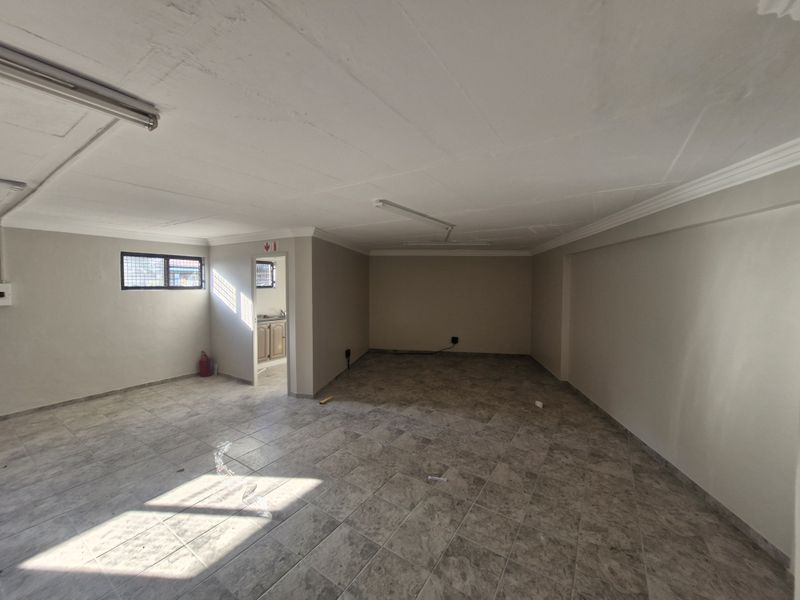 A spacious, empty open-plan commercial area featuring tiled flooring, coved ceilings, and fluorescent lighting, with a small adjacent utility or kitchenette space.