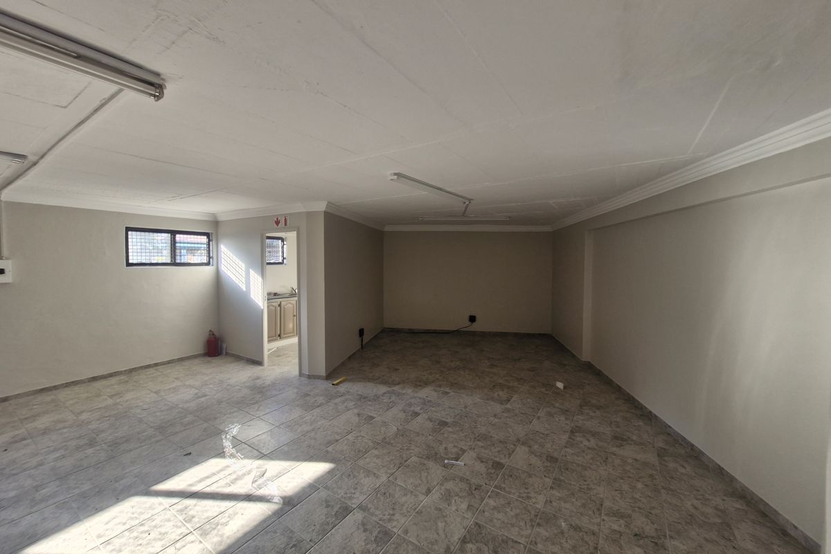 A spacious, empty open-plan commercial area featuring tiled flooring, coved ceilings, and fluorescent lighting, with a small adjacent utility or kitchenette space.