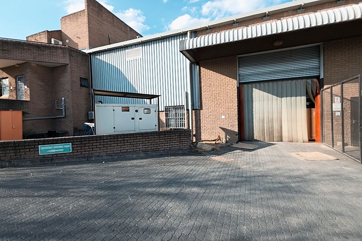 Rear industrial yard featuring a large roller shutter door, brick and metal cladding construction, paved driveway, and external generator unit.