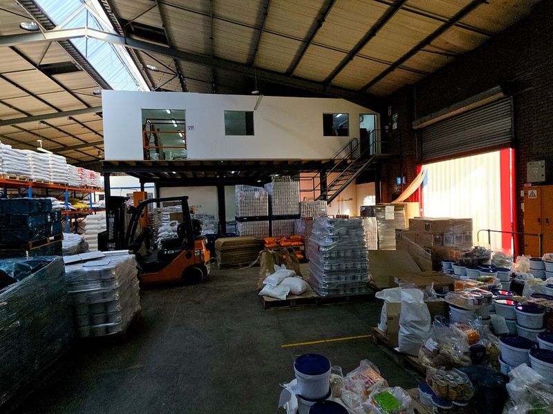 Spacious industrial warehouse featuring a mezzanine level, exposed steel roof trusses with integrated skylights, concrete flooring, and a large roller shutter door.