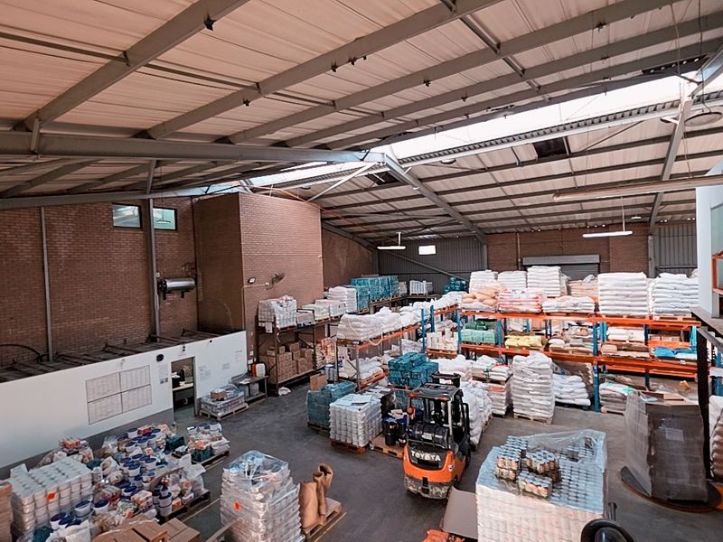 Spacious industrial warehouse featuring high ceilings with exposed steel trusses, concrete flooring, and extensive built-in racking systems for storage.
