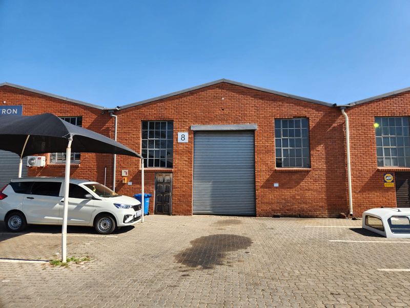 Front elevation of an industrial unit featuring a red brick facade, large roller shutter door, and steel-framed windows with paved parking area.