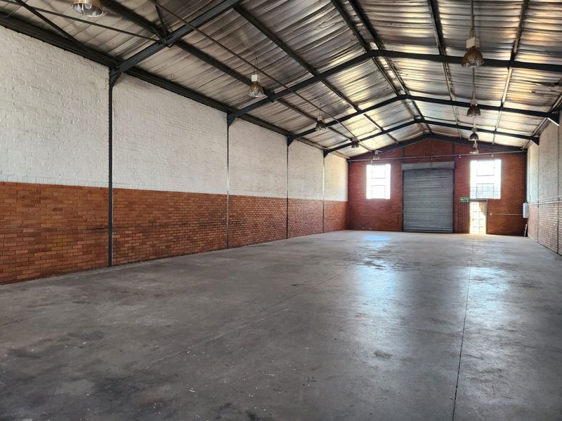 Spacious industrial warehouse unit featuring high ceilings with exposed steel trusses, concrete flooring, brick walls, and a large roller shutter door for easy access.