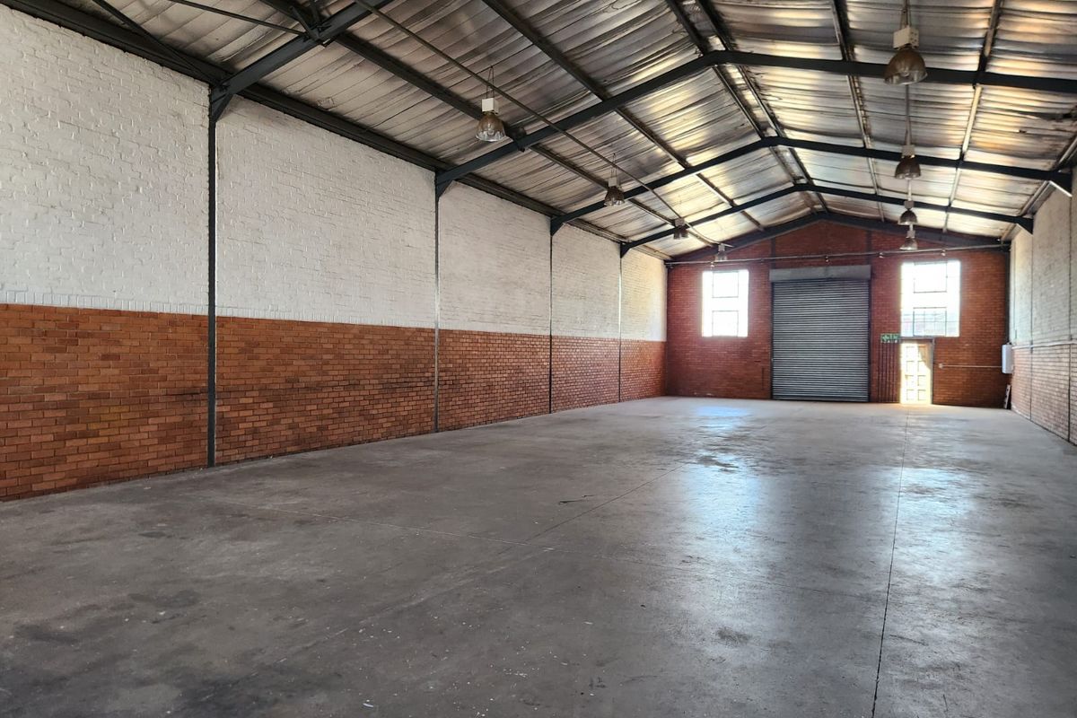 Spacious industrial warehouse unit featuring high ceilings with exposed steel trusses, concrete flooring, brick walls, and a large roller shutter door for easy access.