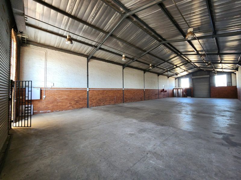 Spacious industrial warehouse unit featuring high ceilings with exposed steel trusses, concrete flooring, brick walls, and a large roller shutter door for easy access.