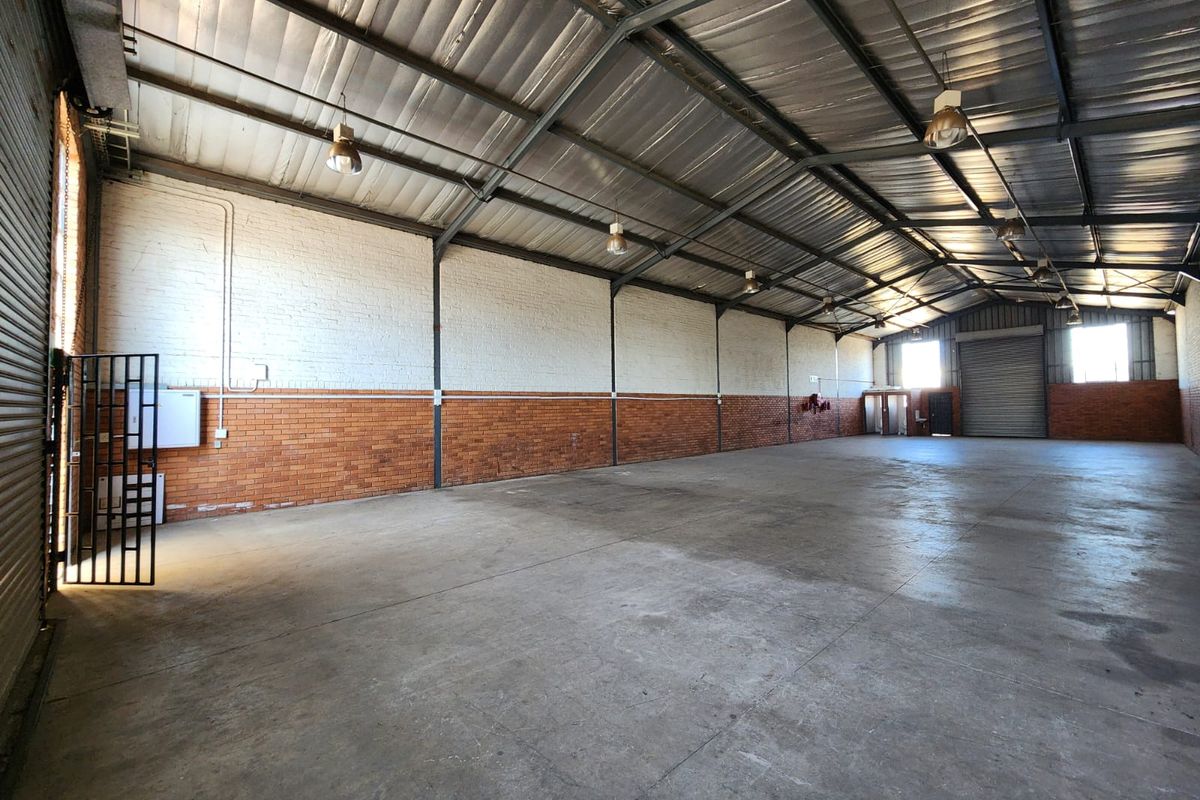 Spacious industrial warehouse unit featuring high ceilings with exposed steel trusses, concrete flooring, brick walls, and a large roller shutter door for easy access.
