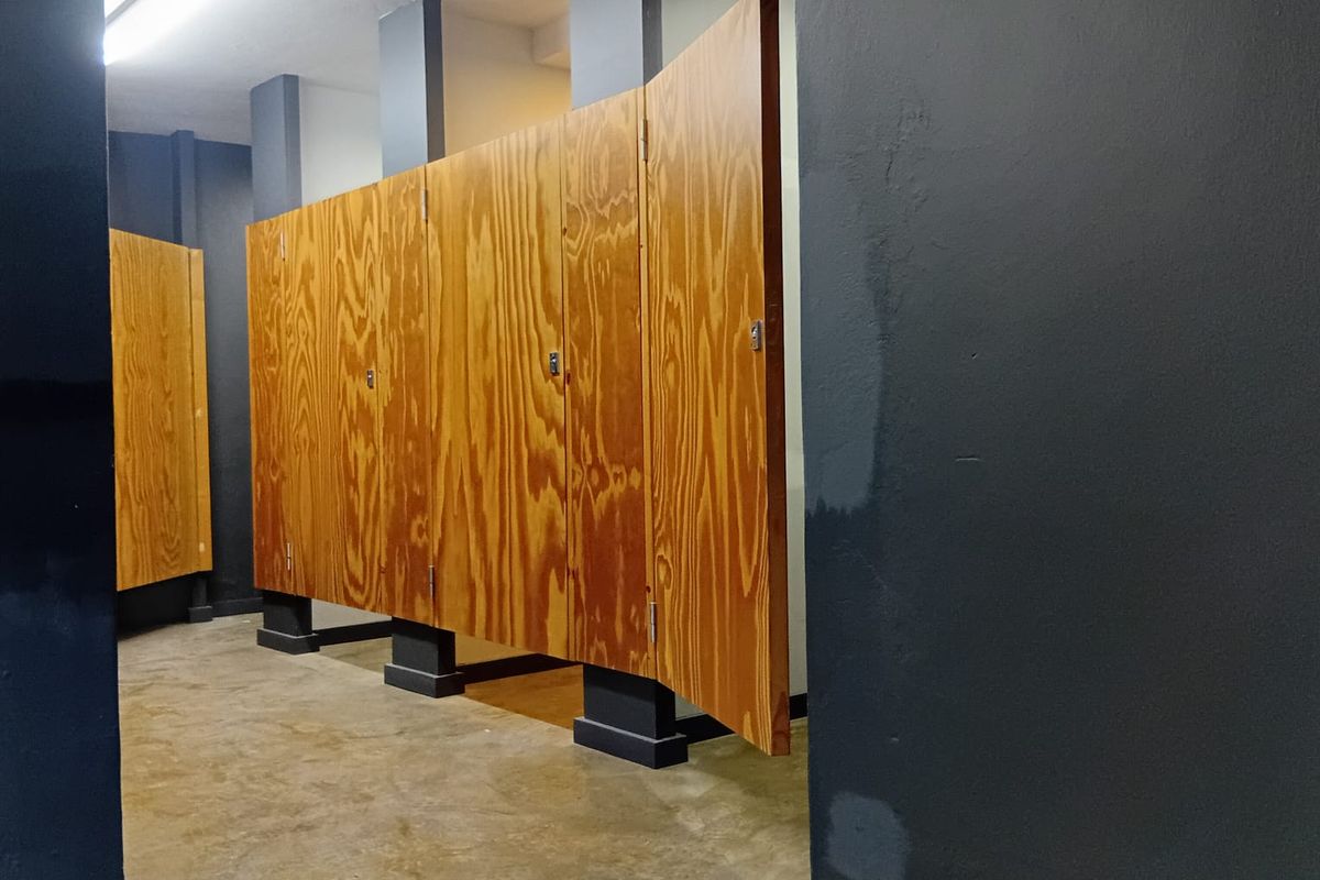 Commercial bathroom facility featuring plywood partitions, concrete flooring, and industrial strip lighting, currently in need of cosmetic updates.