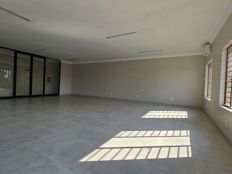 A spacious, empty office area featuring large tiled floors, neutral walls, glass partitioning, and security bars on the windows, providing a clean professional workspace.