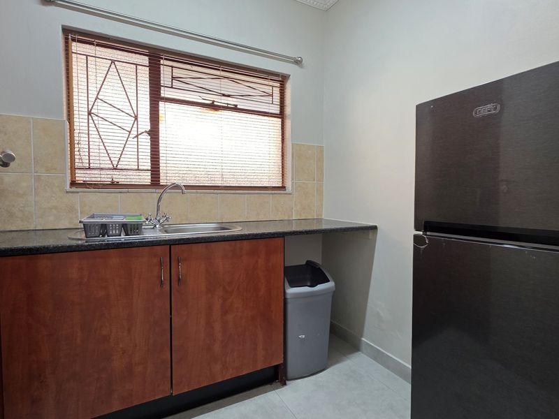 A functional kitchen area featuring built-in wooden cabinetry, granite countertops, a stainless steel sink, and a tiled backsplash under a window with security bars.