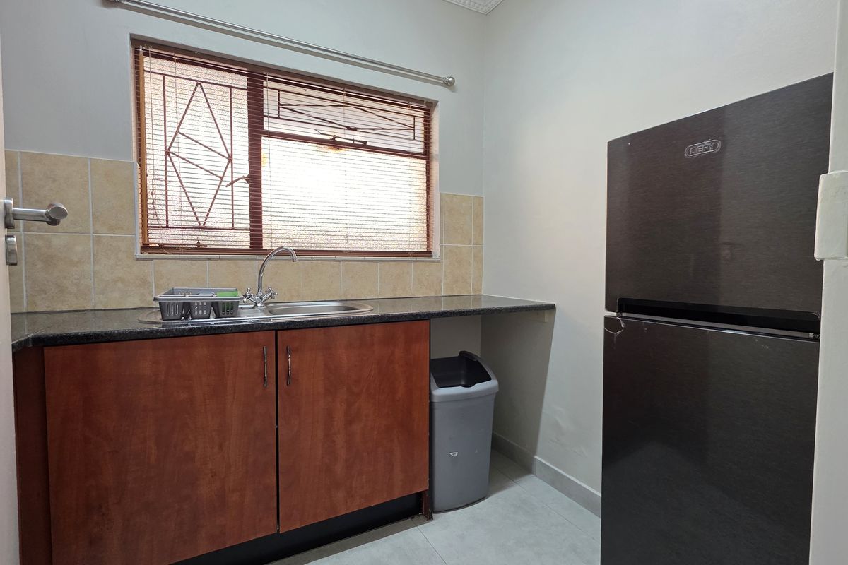A functional kitchen area featuring built-in wooden cabinetry, granite countertops, a stainless steel sink, and a tiled backsplash under a window with security bars.
