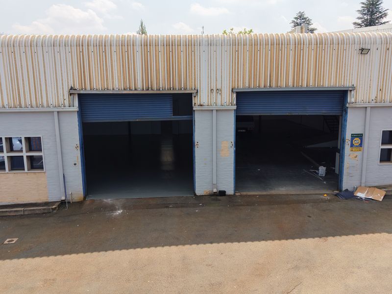 Industrial warehouse exterior featuring dual roller shutter doors, corrugated metal roofing, and multi-pane windows with visible signs of exterior wear.