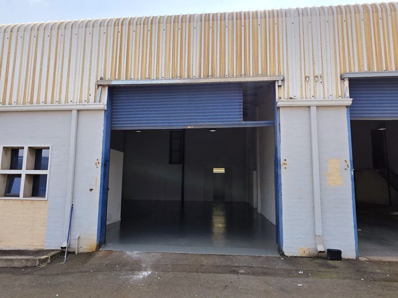 Industrial warehouse exterior featuring corrugated metal cladding, large roller shutter doors, and multiple windows, showing signs of exterior wear and maintenance requirements.