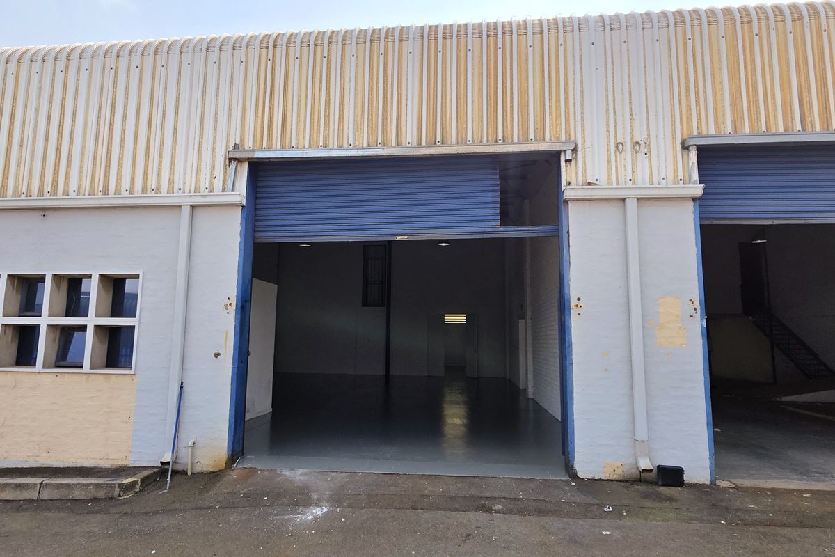 Industrial warehouse exterior featuring corrugated metal cladding, large roller shutter doors, and multiple windows, showing signs of exterior wear and maintenance requirements.