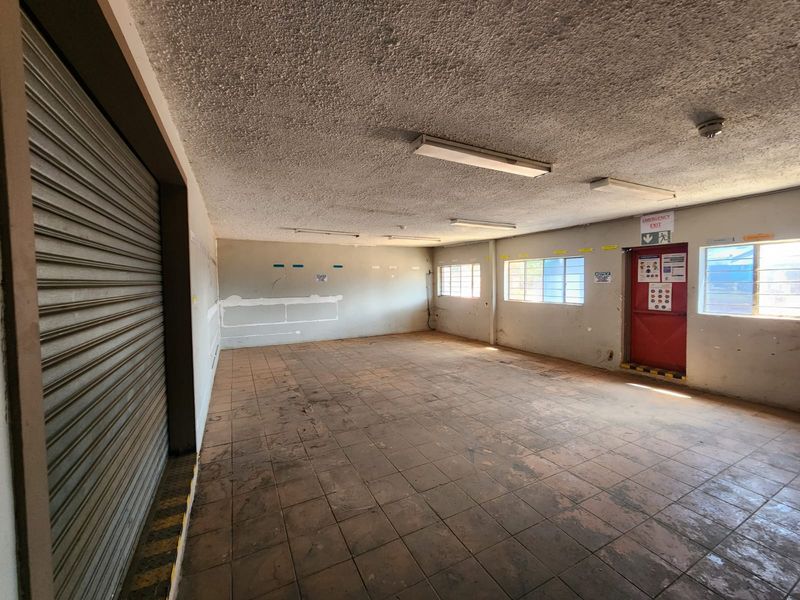 An empty industrial utility space featuring tiled flooring, a large roller shutter door, fluorescent lighting, and a designated emergency exit.