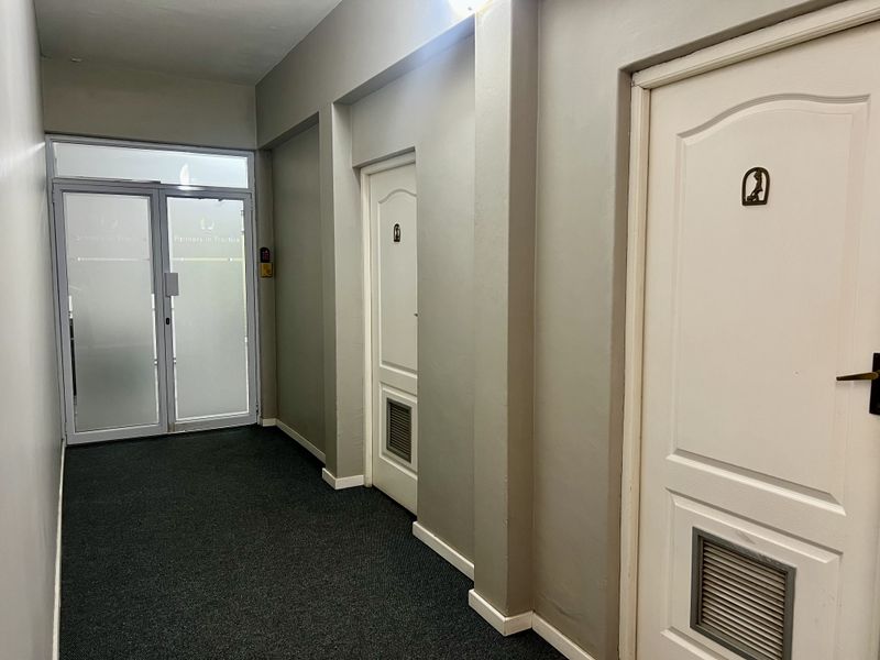 A clean, well-maintained office hallway featuring carpeted flooring, secure glass entrance doors, and multiple internal doors with integrated ventilation grilles.